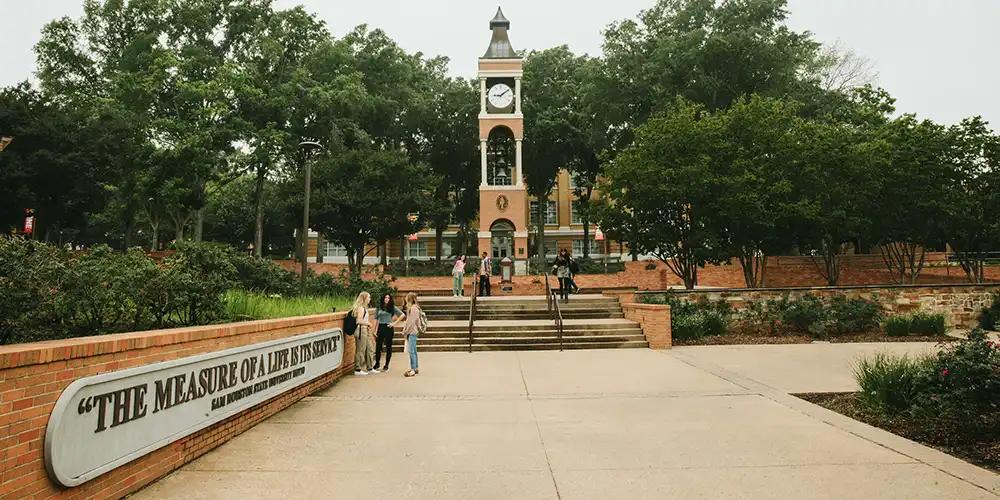 Portrait of the SHSU Bell Tower and the Motto Sign to the Left, with people in photo.