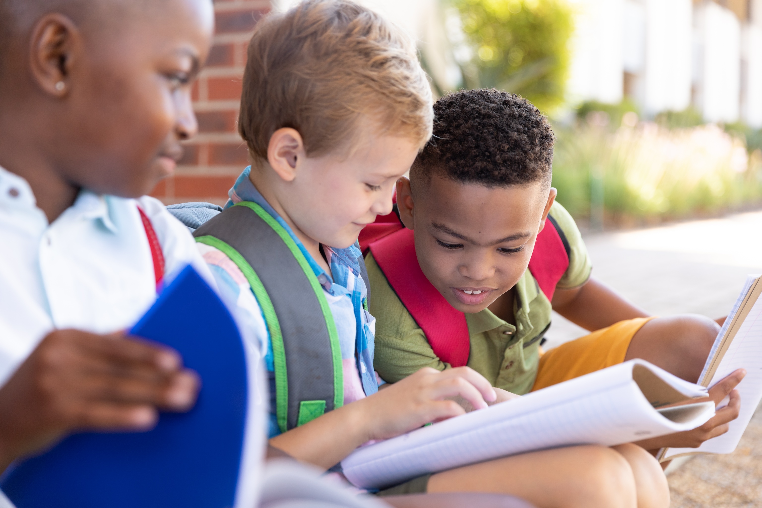 three elementary school age boys wearing backpacks sitting outside looking at a notebook together 