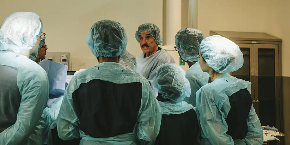 SHSU College of Osteopathic Medicine students in surgical scrubs and caps gather around an instructor during a clinical training session in a medical skills lab.