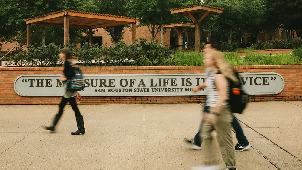 Students walking past SHSU motto sign in plaza 'the measure of the life is its service'.