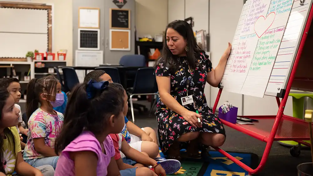 ​​Teacher on her knees, by a whiteboard, taking to her students who are around her sitting down​ 