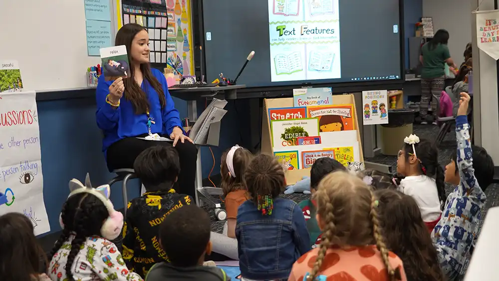 Female teacher seated at the front of the classroom holding up a book for the young students seated on the floor around her