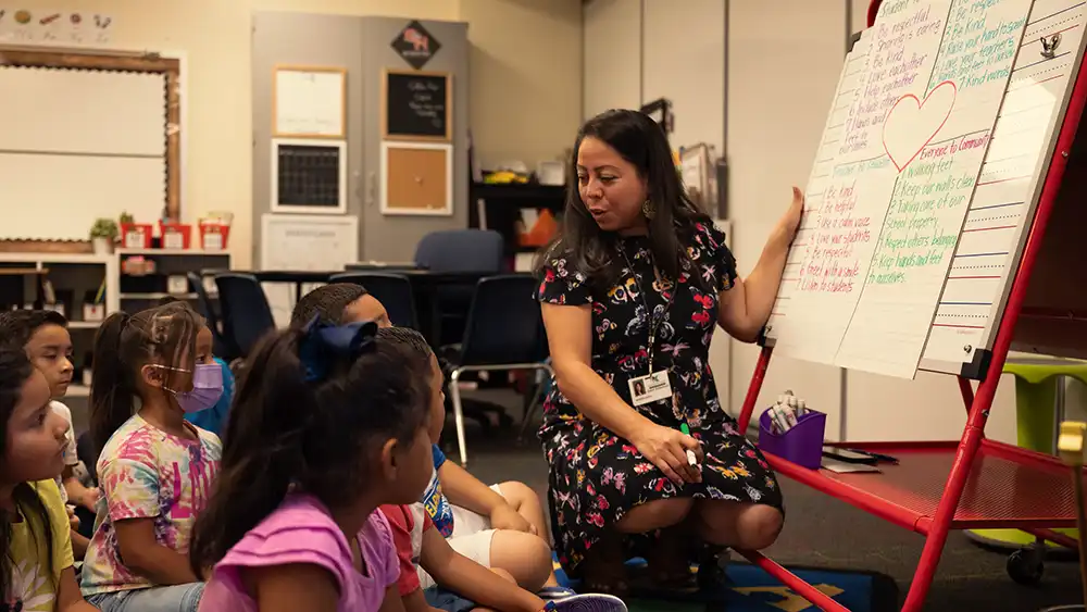 Female teacher kneeling in front of a large dry erase board talking to the young students seated on the floor around her 