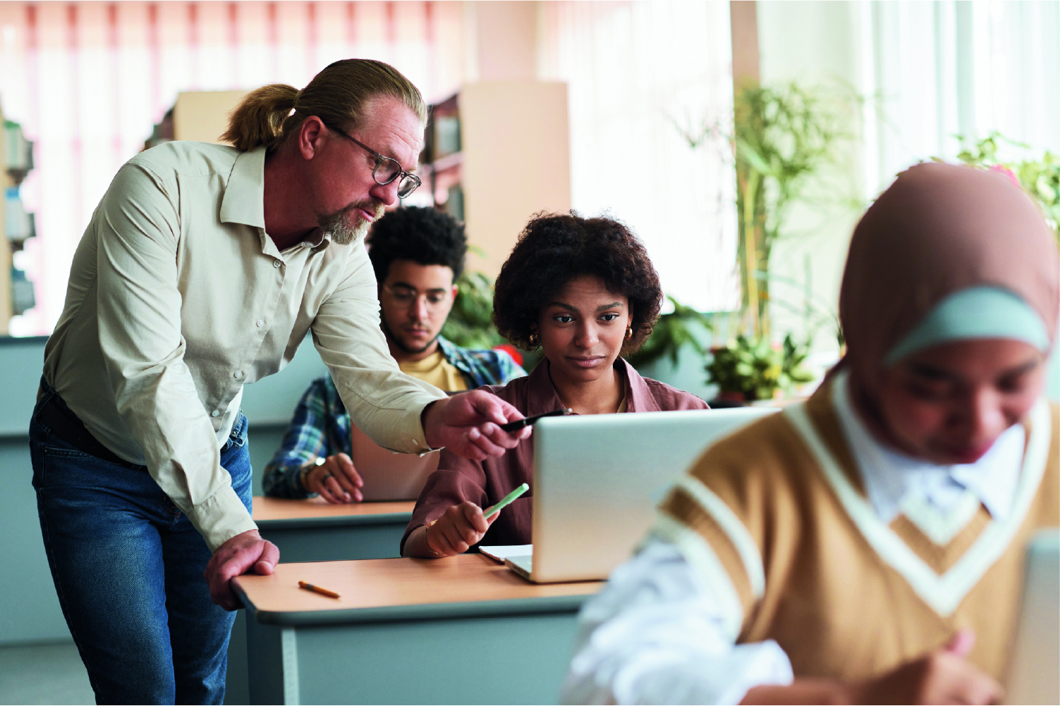 ​​Man in a classroom leaning on student’s desk pointing to her computer screen​ .