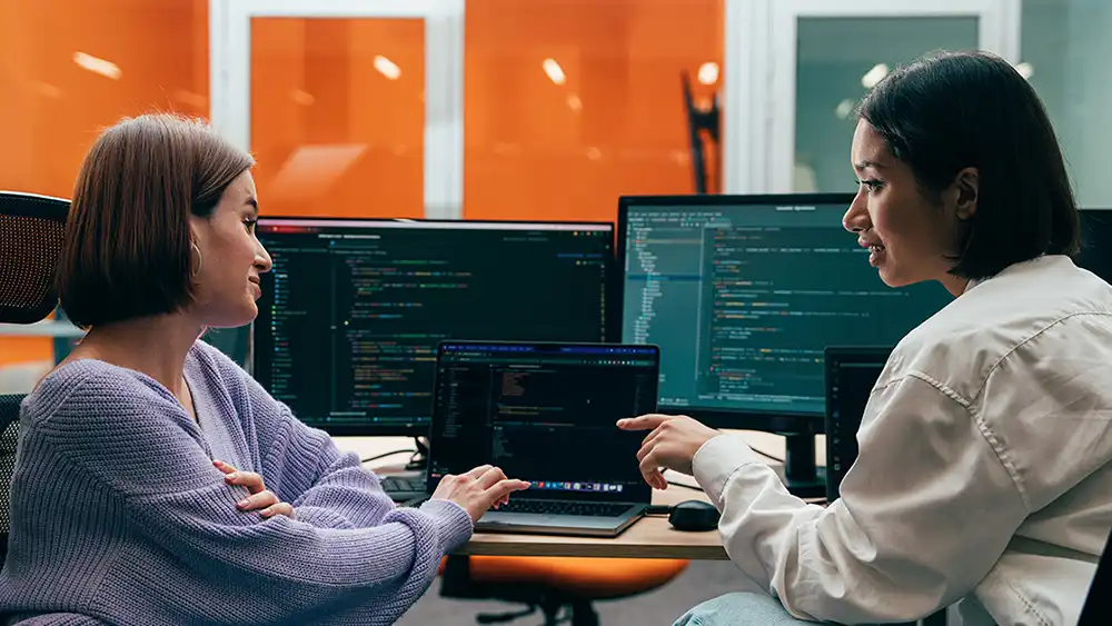 Two people collaborating in front of multiple computer monitors displaying code in a modern office setting.
