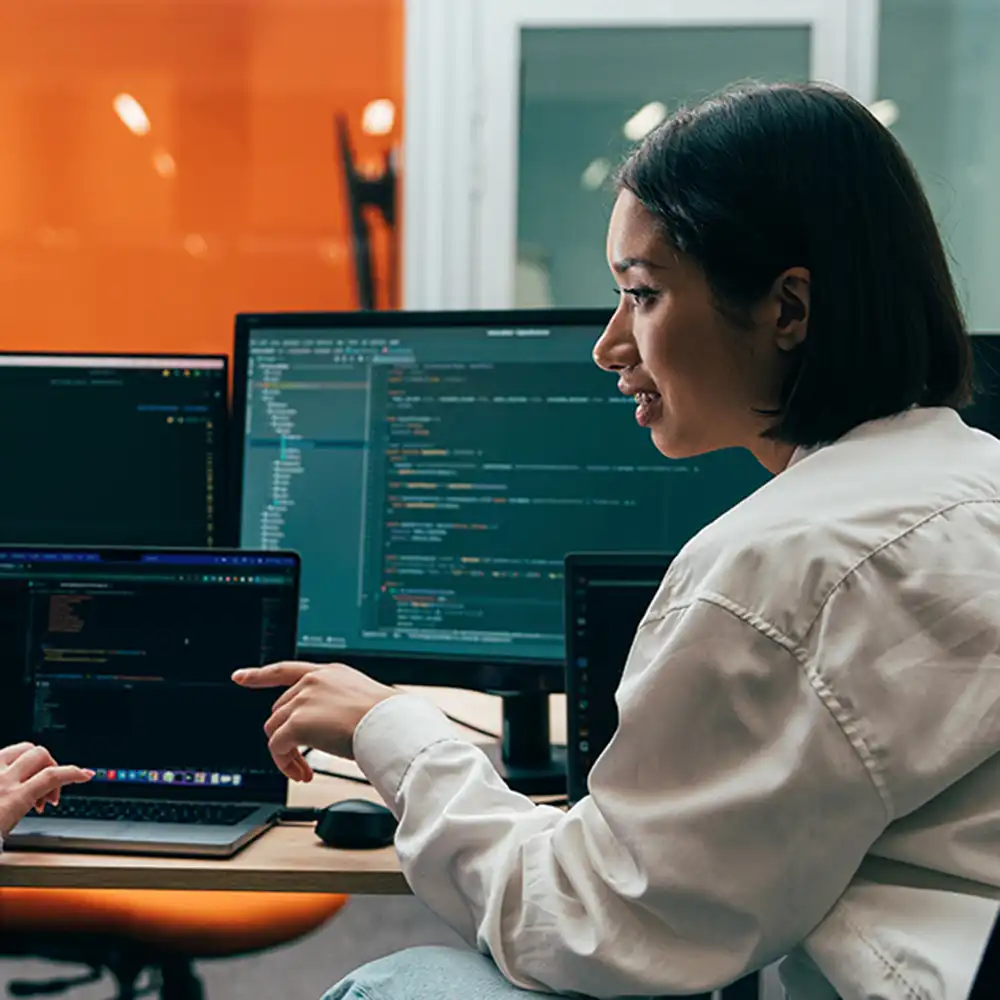 Person pointing at code displayed on multiple computer monitors while collaborating in a modern office setting.