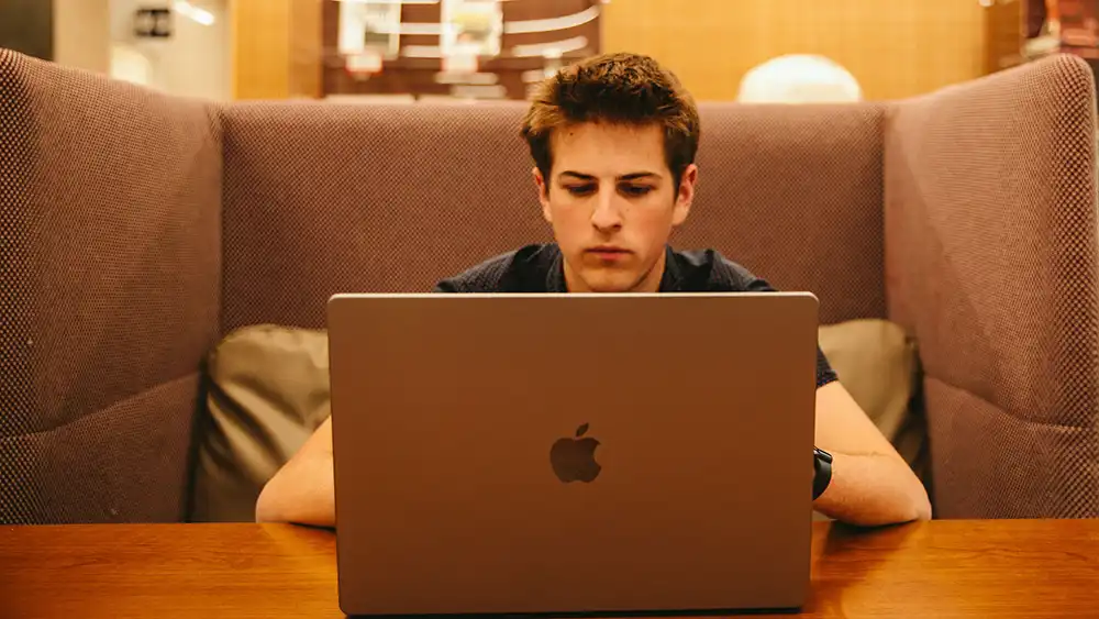 ​​A college student sits in a booth focused on a laptop, reviewing information on the screen. The setting appears to be a quiet study area or lounge.​ 