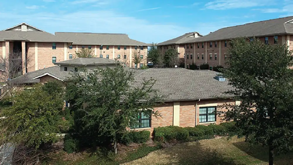 ​​Exterior view of Bearkat Village Apartments at Sam Houston State University. In the foreground is a single-story clubhouse with a grey roof and large windows, surrounded by neatly trimmed bushes and trees. Behind the clubhouse, three multi-story residential buildings with light brown brick exteriors and grey roofs are visible. The sky is clear and blue with a few wispy clouds.​ 