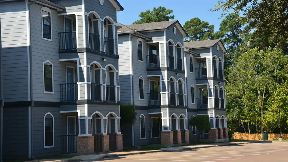 Exterior view of Campus Edge Apartments at Sam Houston State University. The three-story buildings have a gray and white color scheme, with balconies featuring black railings. The architecture includes arched windows and doorways on the ground floor. Trees surround the community, and a paved area is visible in front of the buildings. 