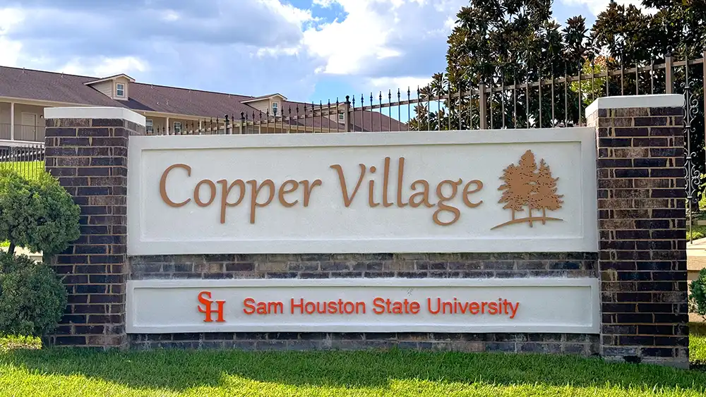 ​​Exterior view of Copper Village Apartments at Sam Houston State University. A brick and stone sign in the foreground displays 'Copper Village' in large copper-colored letters on a white background, with an illustration of two trees to the right. Below, 'Sam Houston State University' is written in smaller red letters next to the university's orange 'SH' logo. In the background, a residential building with beige siding and a brown roof is partially visible behind a wrought iron fence and trees. The sky is partly cloudy.​ 
