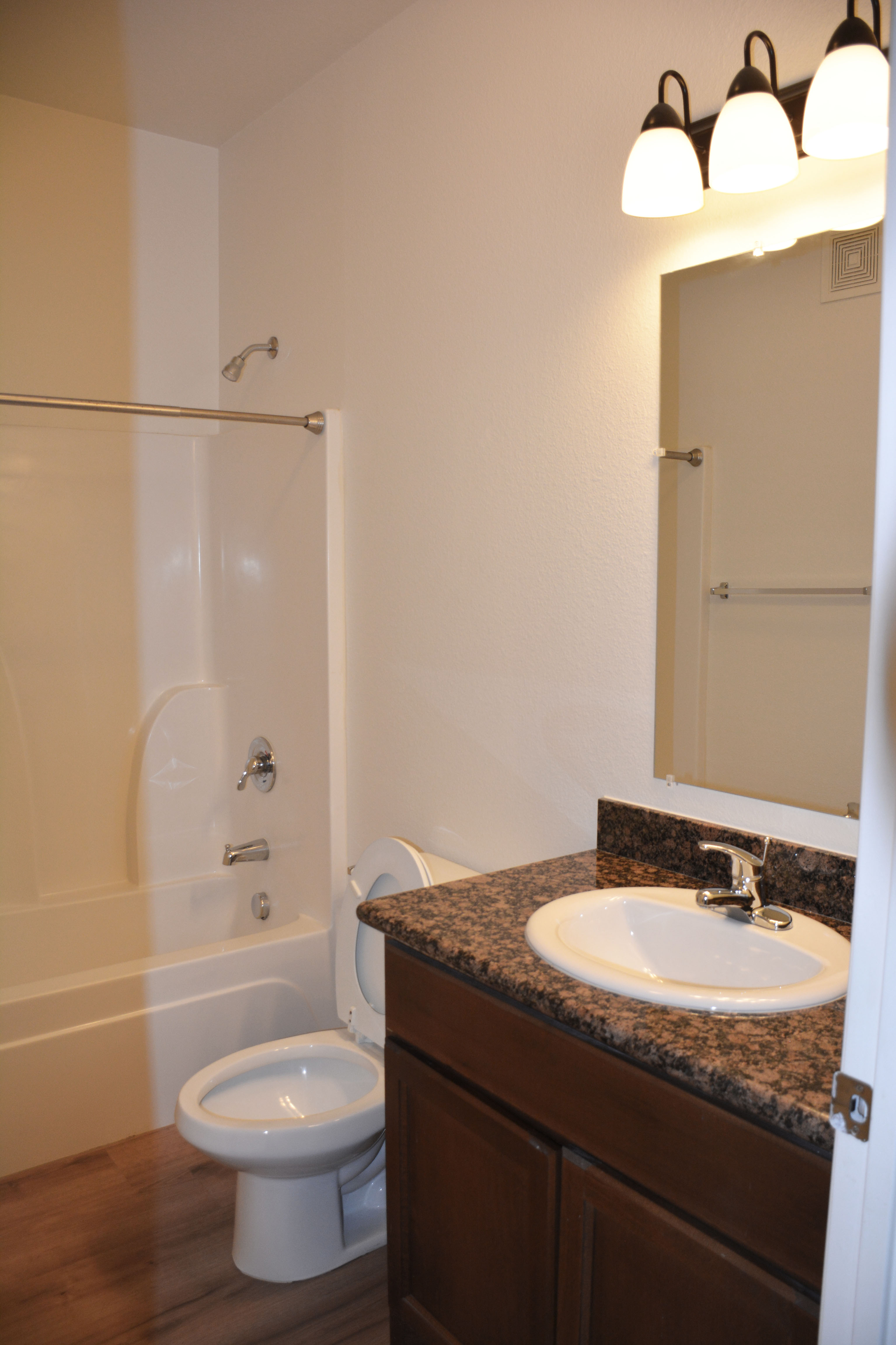 Bathroom in SHSU’s Copper Village Apartments with a white bathtub and shower, toilet, and sink with granite countertop. A large mirror and three light fixtures are mounted above the sink. The room has wood-style vinyl flooring and white walls.