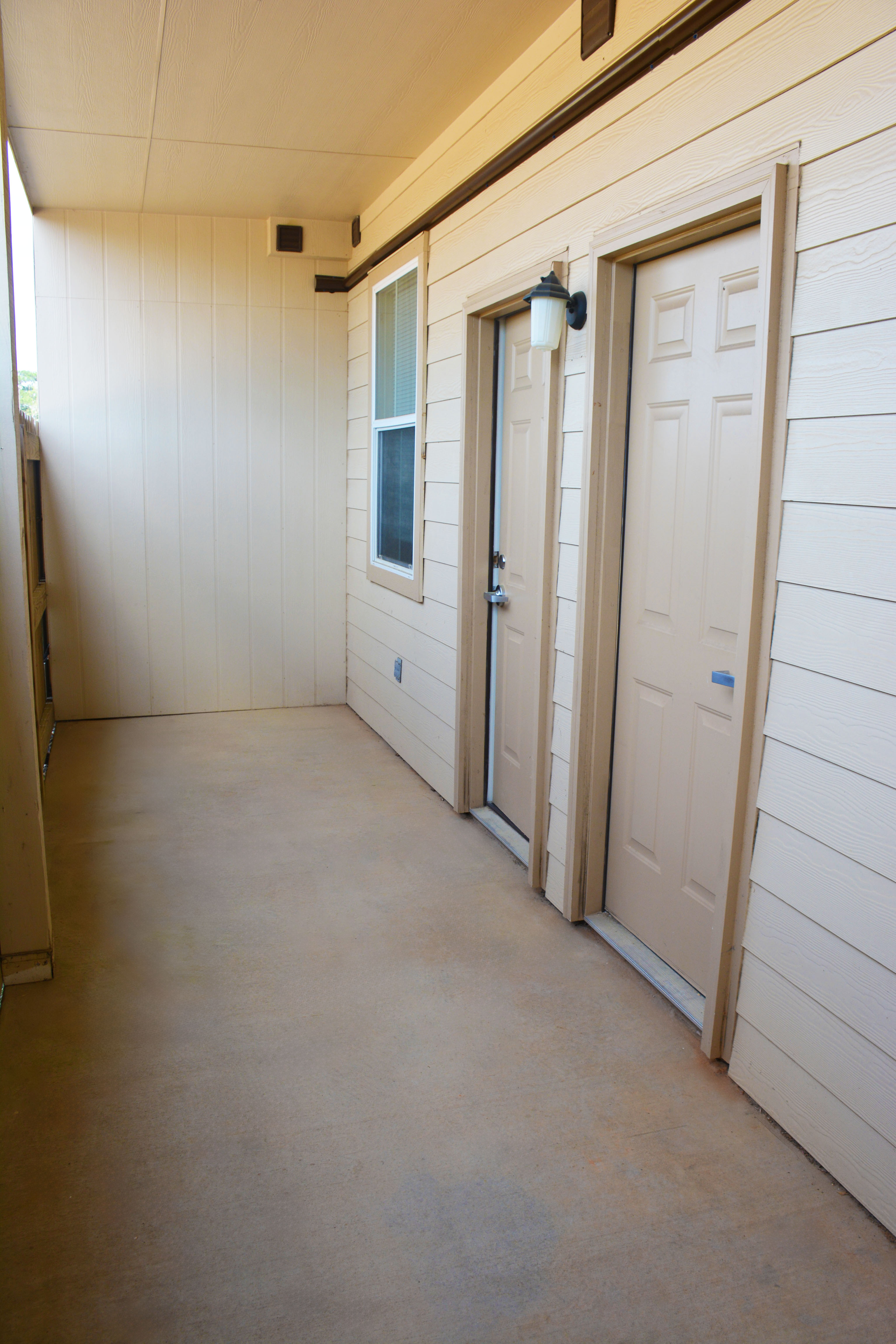 Patio at SHSU’s Copper Village Apartments with beige concrete flooring, cream-colored paneled walls, two identical doors with small windows, a wall-mounted light fixture, and a single window with white trim. Natural light enters from the open side.
