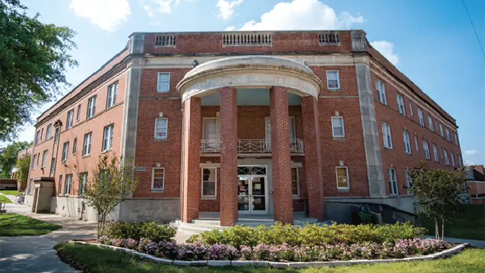 Front view of Elliott Hall at Sam Houston State University, a historic red-brick, all-female residence hall featuring large columns, arched entryway, and landscaped flower beds.  