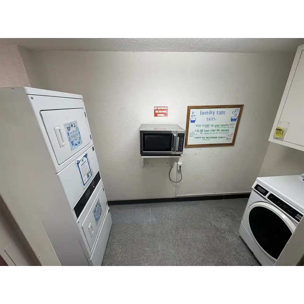 Laundry room in SHSU’s Estill Hall with stacked white washers and dryers, tiled floor, white walls, a desk with a blue chair, and a wall-mounted sink in the corner. Fluorescent lights illuminate the space.