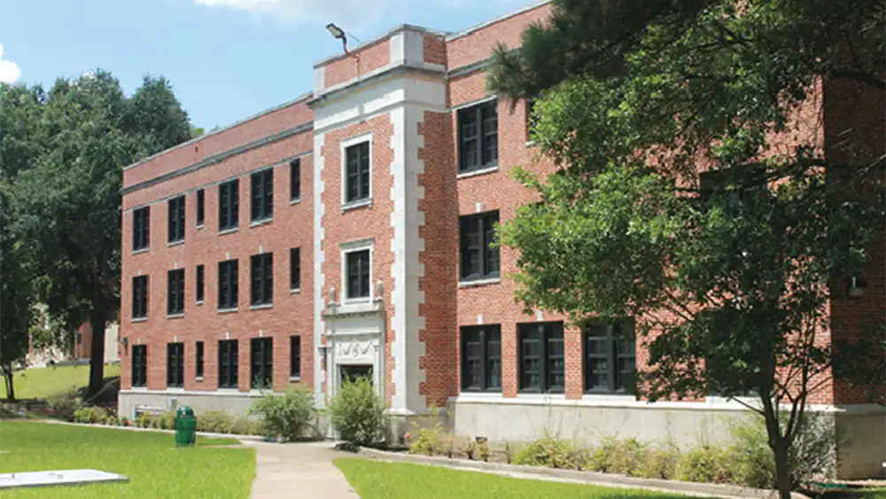 Three-story red brick residence hall with black-framed windows and a central entrance framed by white stone. The building is surrounded by trees, shrubs, and a grassy lawn with a sidewalk leading to the entrance.  