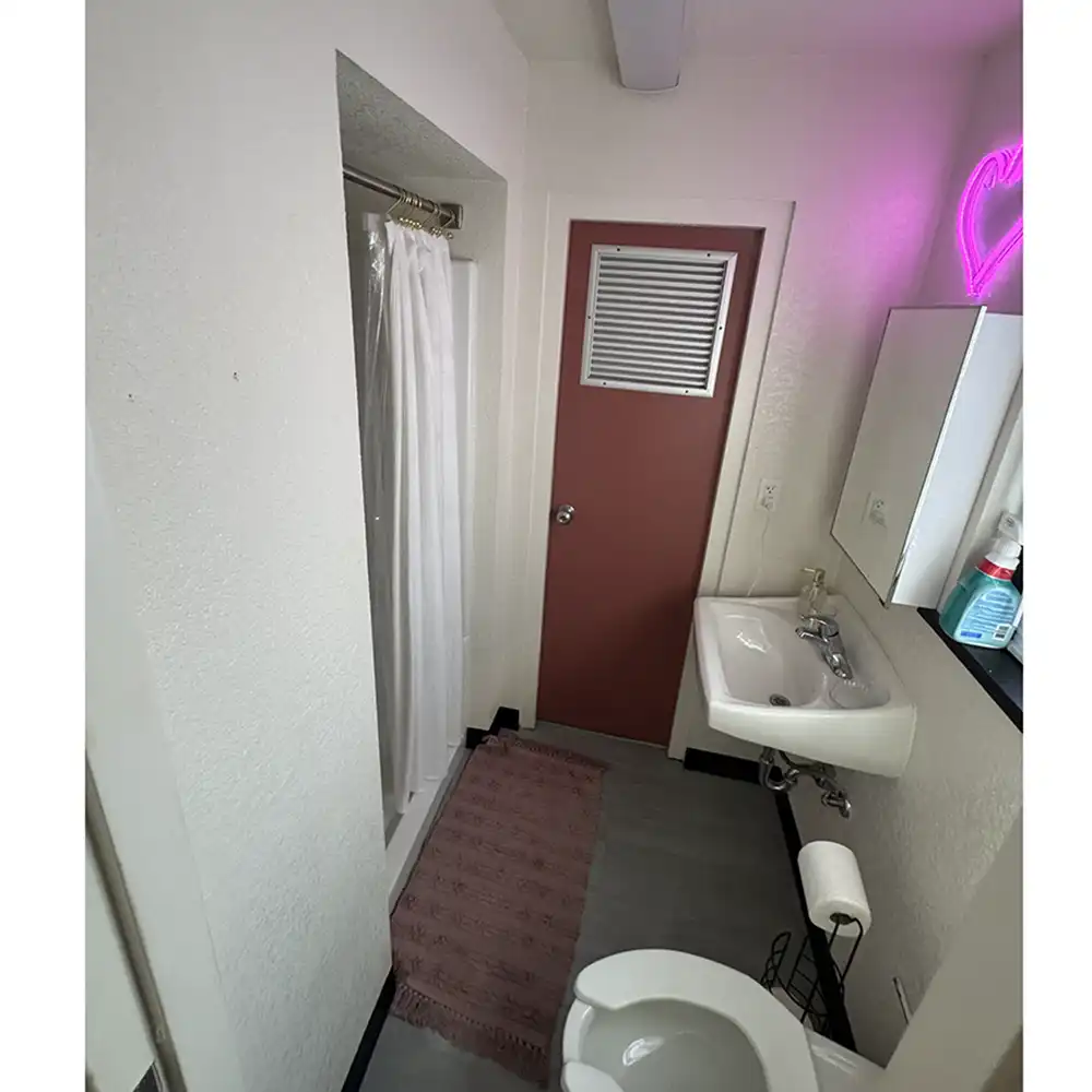 Bathroom in SHSU’s Jackson-Shaver Hall with a shower and white curtain, a pink door with a vent, and a white sink with a mirrored cabinet above. A pink rug lies on the floor, and a neon heart-shaped light glows above the sink area.