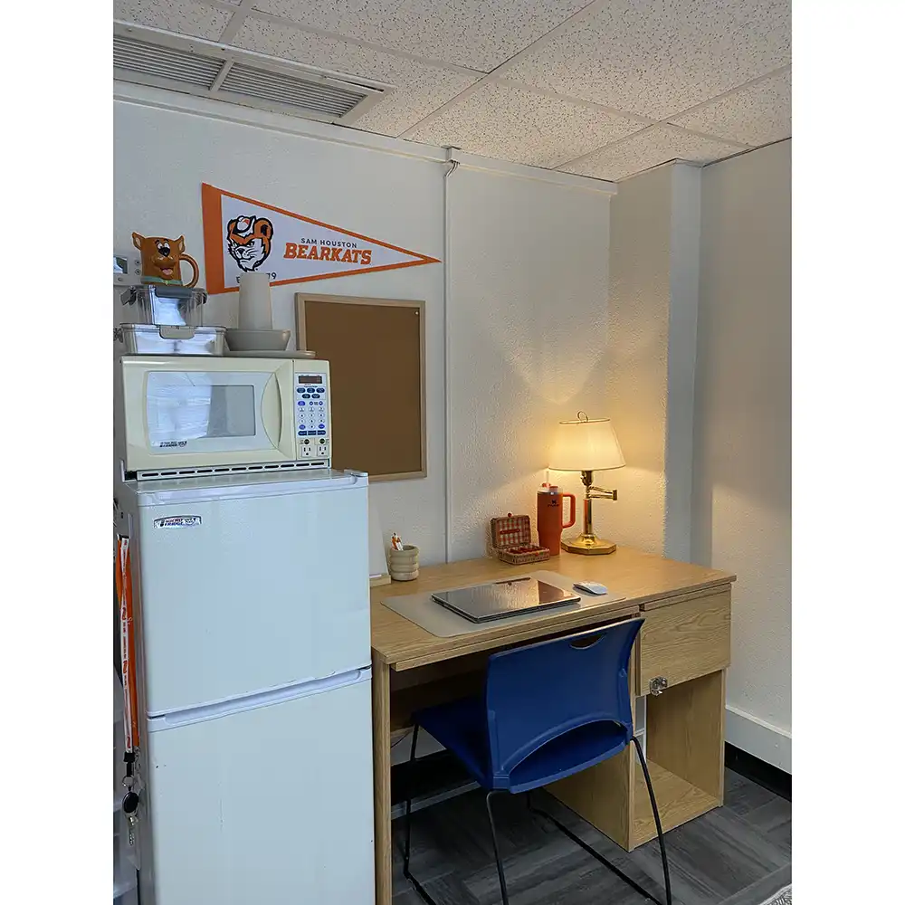 Corner of a bedroom in SHSU’s Jackson-Shaver Hall featuring a wooden desk with a blue chair, laptop, and lamp. A mini fridge with a microwave on top sits beside the desk. Above are an orange 'Sam Houston State BEARKATS' pennant and an empty corkboard. The room has white walls and ceiling tiles.