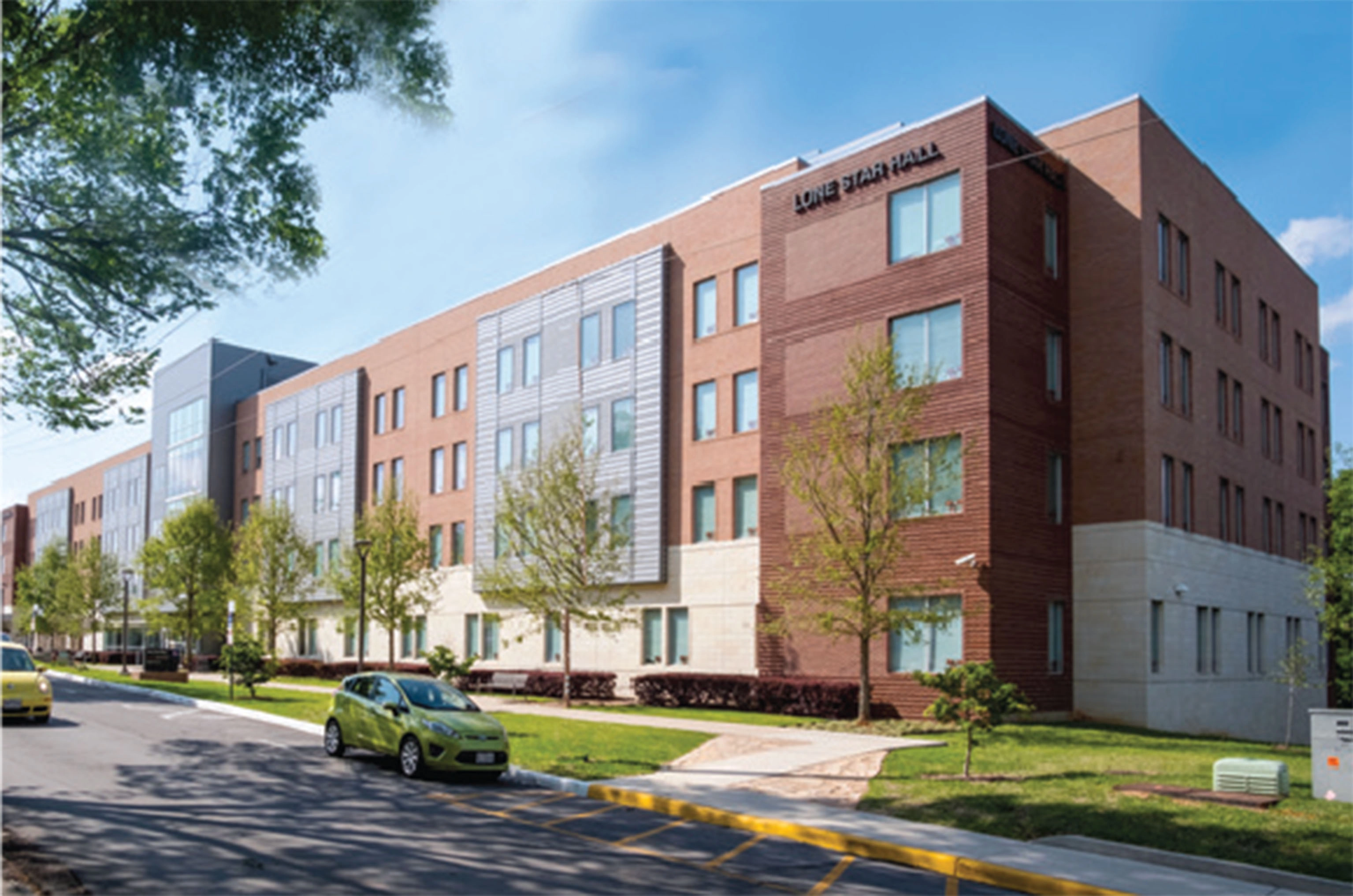​​Modern four-story residence hall named "Lone Star Hall," featuring a mix of red brick, light stone, and silver metal paneling. The building is lined with large windows and surrounded by young trees and a landscaped lawn, with cars parked along the street in front.​ 