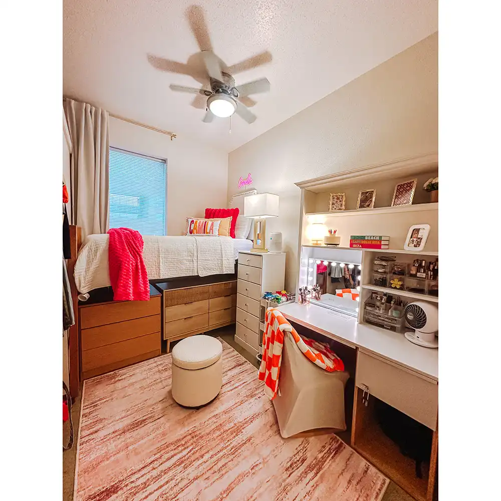 Bedroom in SHSU’s Lone Star Hall with a lofted bed featuring white bedding, red accents, and four plush pillows.  Wooden drawers are beneath the bed, and a tall dresser stands nearby. A white desk with shelves holds books, photos, and makeup items, with a chair draped in an orange and white checkered blanket. The room includes a pink rug, sheer curtains, and a ceiling fan light.