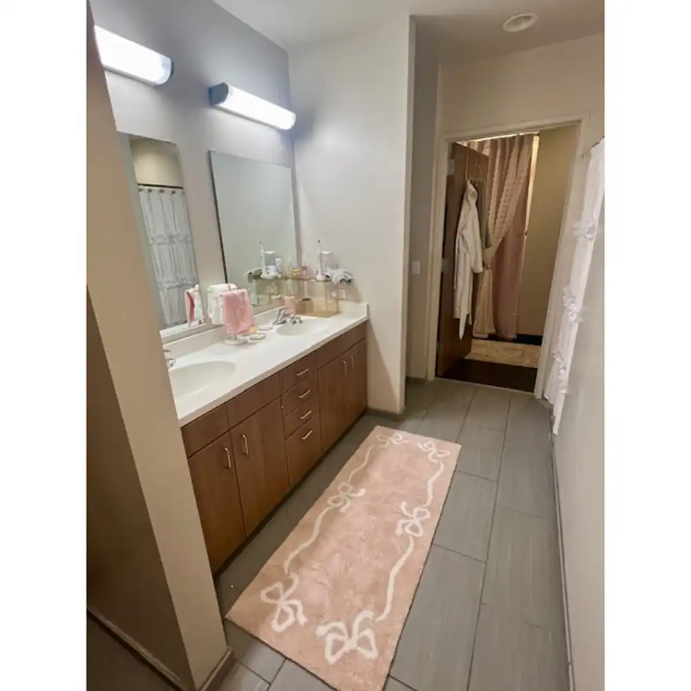 A Piney Woods Hall vanity area at SHSU with a long countertop, double sinks, and large mirrors under bright wall-mounted lights. The wooden cabinets below provide storage, while a pink rug with white bow designs runs along the tiled floor. Towels, toiletries, and decor items sit neatly on the counter, and a doorway in the back reveals hanging robes and curtains.