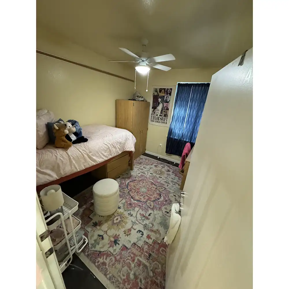 Bedroom in SHSU's Raven Village featuring a single bed with a pink comforter and stuffed animals, storage drawers underneath, and a wooden wardrobe beside it. A ceiling fan with lights hangs overhead. On the right, a window with dark curtains sits above a desk and chair. The floor has a colorful patterned rug, with an ottoman near the bed’s foot. A small white shelving unit with personal items is placed near the open door.