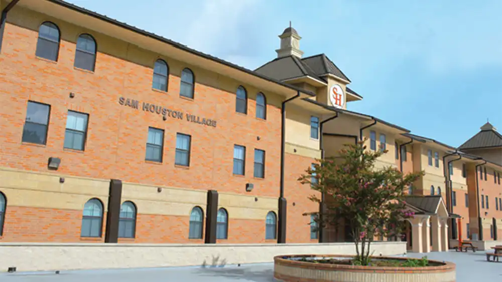 Image Alt Text   ​​Exterior view of Sam Houston Village at Sam Houston State University. The three-story residential building features red brick walls with beige stone accents and multiple windows, including arched ones on the ground floor. 'SAM HOUSTON VILLAGE' is displayed in large letters on the side of the building. A small courtyard in front includes a circular brick planter with a tree and surrounding greenery. The sky is mostly clear with a few clouds.​ 