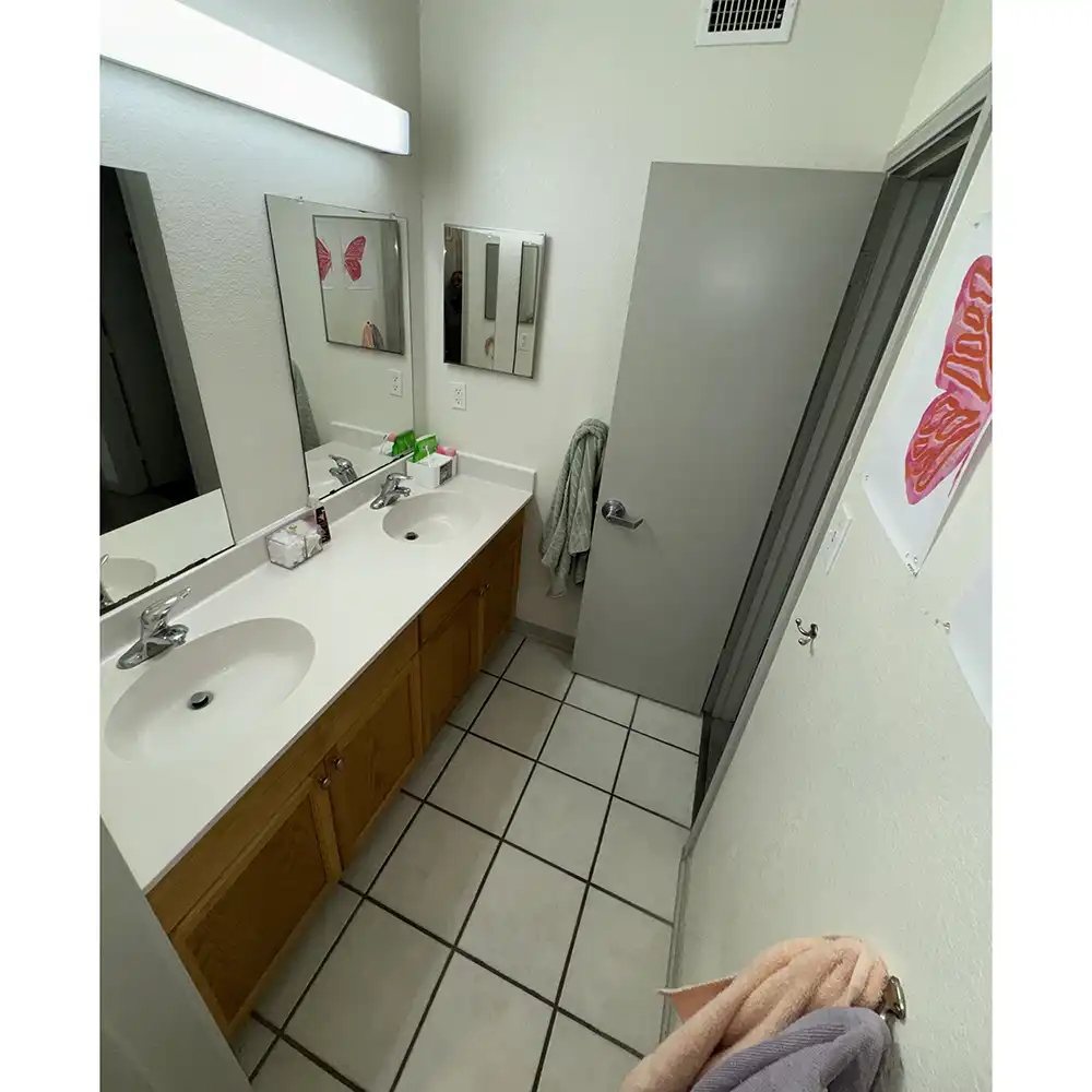 Entry area of a bathroom at SHSU's Sam Houston Village featuring a double sink vanity with a white countertop and two mirrors above each sink. Toiletries like soap dispensers and toothbrush holders are on the counter. A towel hangs on the wall near a slightly open door, and a pink butterfly decoration is visible in the mirror's reflection. The floor is tiled with white square tiles, and additional towels hang on hooks near the entrance.