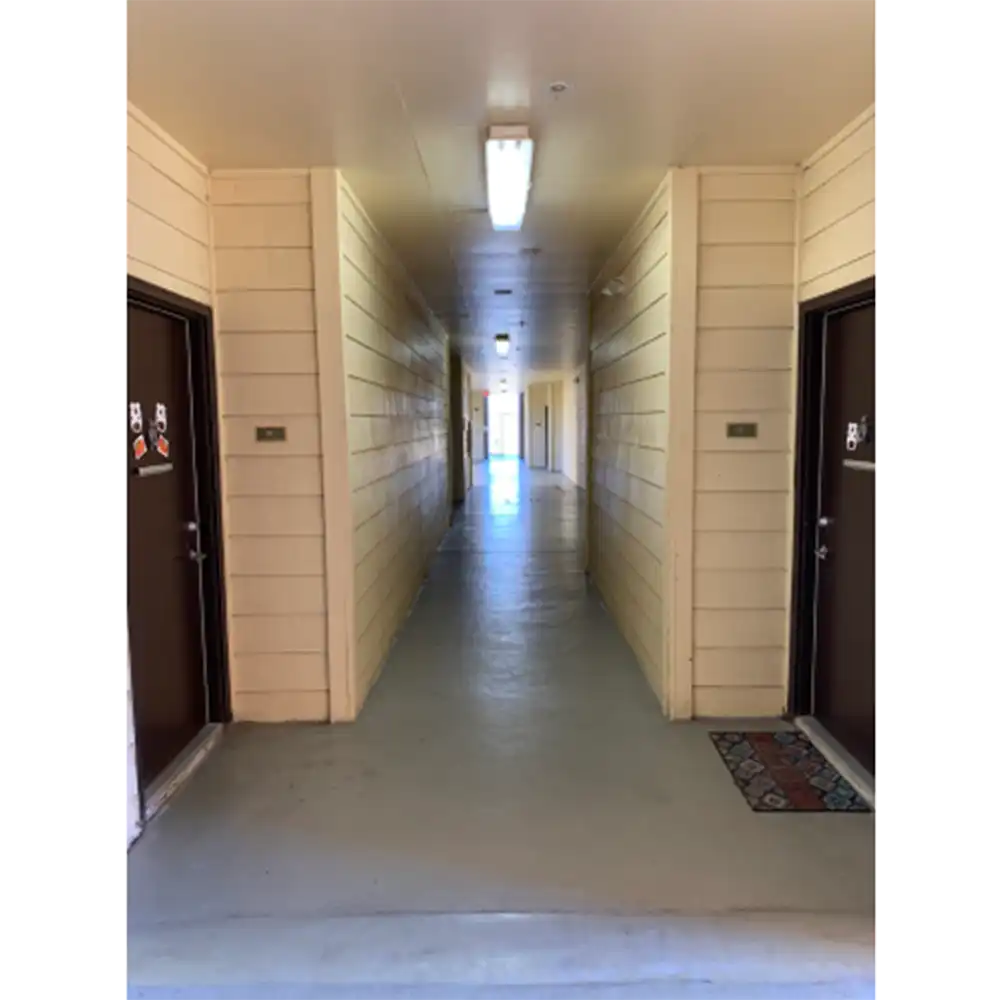 Breezeway at Sam Houston Village, SHSU, featuring a long corridor with beige wooden walls, a smooth concrete floor, and brown doors on both sides with room numbers and small decorations. Fluorescent lights are evenly spaced along the ceiling, and natural light is visible at the far end from an open area or window.