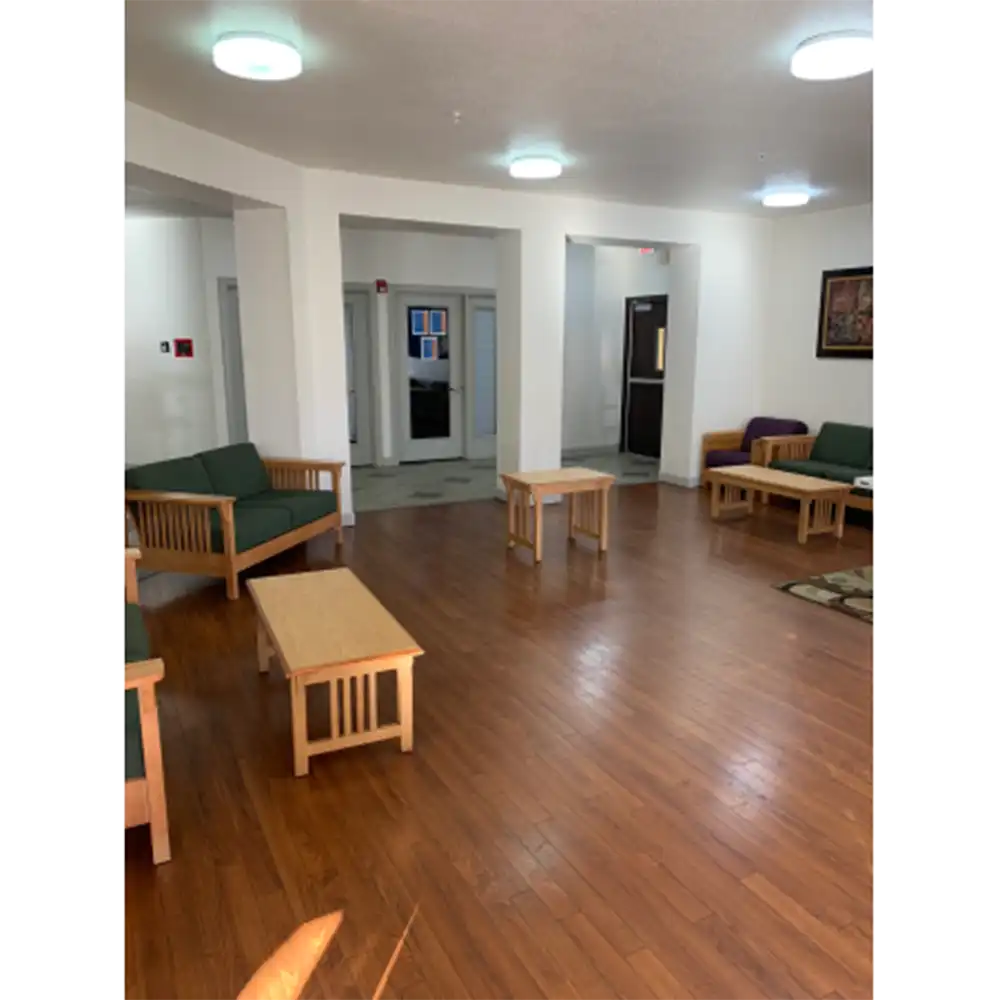 Lobby of Sam Houston Village at SHSU featuring wooden flooring, several wooden tables and chairs with green cushions, and three seating areas with couches or chairs and coffee tables. The white walls are lit by multiple ceiling lights, and doorways lead to other parts of the building including a glass entrance and a hallway. A framed picture is hung on the right wall.