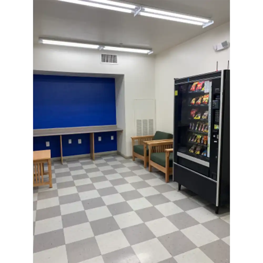 Room at Sam Houston Village, SHSU, featuring a snack-filled vending machine on the right, two wooden chairs with green cushions and a small table on the left, a blue accent wall with a long counter and electrical outlets in the background, and a checkered floor of light and dark gray tiles under fluorescent ceiling lights. 