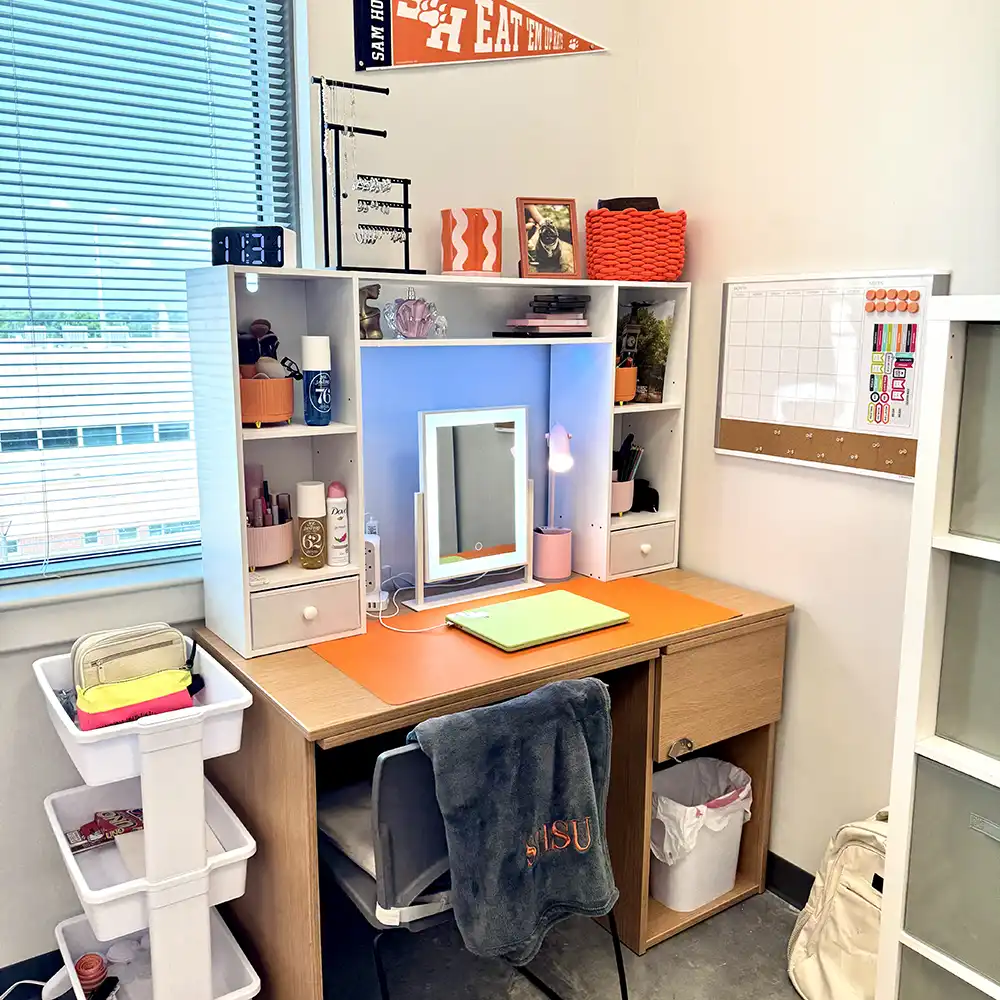 Desk and chair in SHSU’s San Jacinto Hall featuring a wooden desk with orange desktop cover, white shelving unit with storage cubbies, a mirror with lights, study supplies, and a chair with an SHSU blanket draped over it. A window, wall calendar, and Sam Houston pennant are visible in the background.