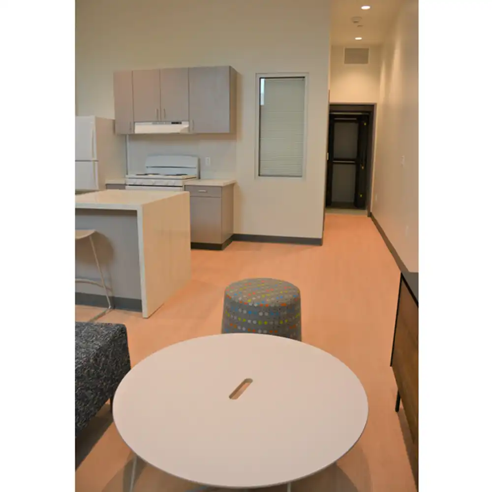 Living room of SHSU's San Jacinto Studio Apartments featuring modern design with light-colored walls and flooring. Visible furnishings include a round white coffee table with a center slot, a gray textured sofa, and a colorful polka-dot ottoman. In the background, an open kitchen area has light wood cabinets, a white refrigerator, electric stove, and a small island counter with two bar stools. A hallway is visible on the right side leading to another part of the apartment.