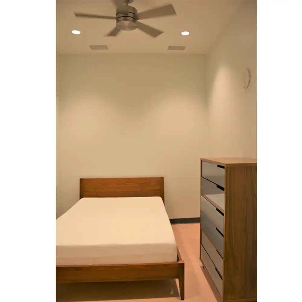 Bedroom in SHSU's San Jacinto Studio Apartments featuring a wooden bed frame with a mattress against the back wall. A tall wooden dresser with multiple drawers is positioned to the right of the bed. The room includes recessed ceiling lights, a modern four-blade ceiling fan, light-colored walls, and laminate flooring.