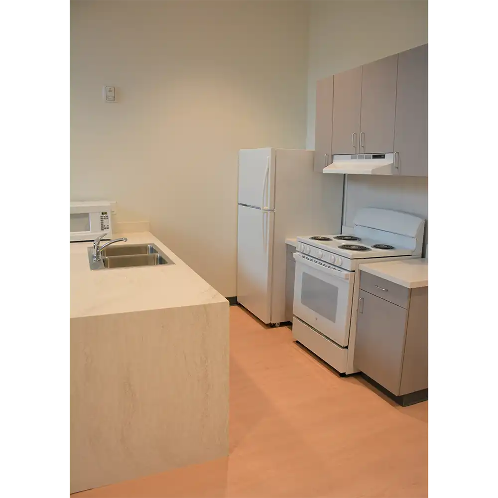 Kitchen area of SHSU's San Jacinto Studio Apartments featuring a white refrigerator, white electric stove with oven, and a microwave on the countertop. A stainless steel sink is set into a marble countertop, with light gray cabinets above and below. The flooring is light-colored laminate.