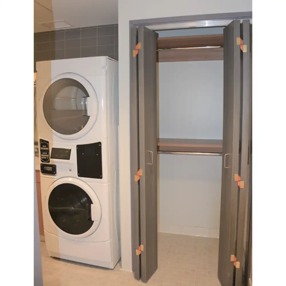 Washer and dryer area in SHSU's San Jacinto Studio Apartments featuring a stacked white unit with control panels in between. Next to it is an open closet with double doors, containing a wooden shelf and hanging rod. The floor is tiled, and the walls are painted light gray