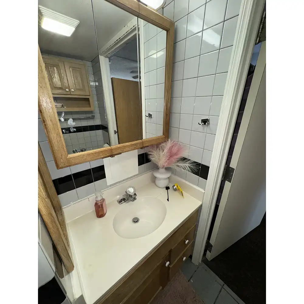 Bathroom in SHSU’s White Hall with a white sink and wooden cabinet, a large mirror with a wooden frame, and white tiled walls with a black horizontal stripe. The countertop holds a pink soap dispenser and a white vase with pink and beige decorative feathers. A wooden door and shelving are visible in the mirror’s reflection.