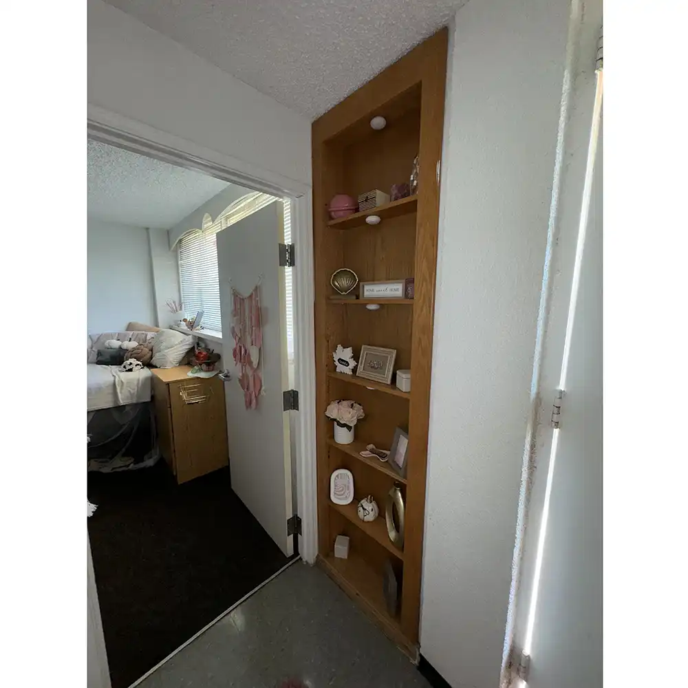 Built-in wooden bookshelf in SHSU’s White Hall with five decorated shelves holding framed photos, candles, a pink flower arrangement, a mirror, and inspirational signs. An open door in the background reveals more room decor.