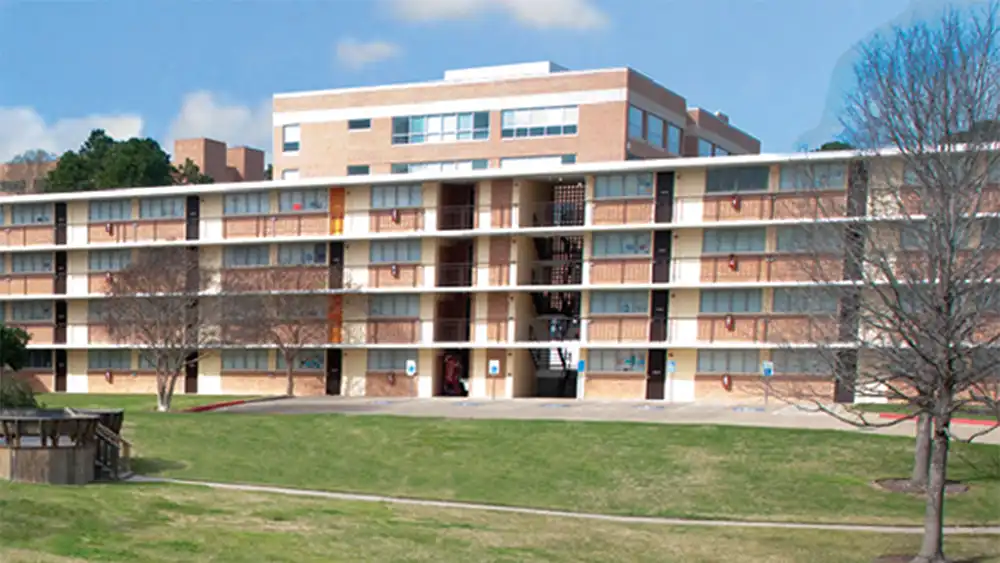 Exterior of SHSU’s White Hall, a multi-story building with brick and concrete facade, large windows, and balconies. Trees and a green lawn are in front, with a clear blue sky overhead.