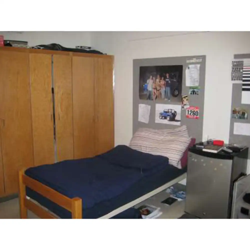 Bedroom in one of SHSU's Small Houses featuring a single bed with a blue comforter and striped pillow, a bulletin board with photos and papers above the bed, a small refrigerator with personal items on top, and a large wooden wardrobe on the left. The decor is simple and functional, reflecting typical student housing