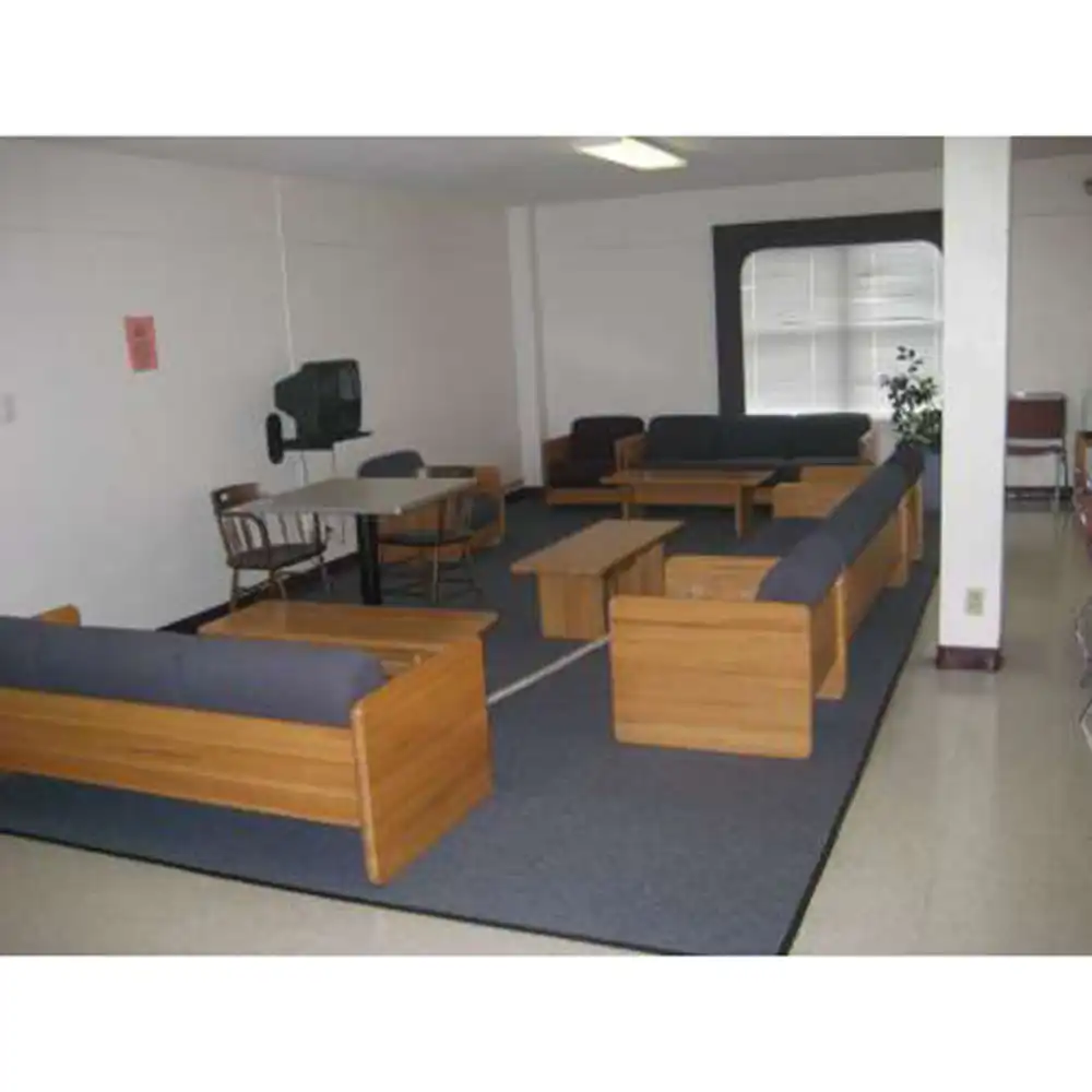 Lobby in one of SHSU's Small Houses featuring wooden-framed couches and chairs with dark blue cushions, several coffee tables, a small round table with two chairs near a wall-mounted TV, a large blue rug, white walls, fluorescent ceiling lights, and a window with natural light and a potted plant nearby.