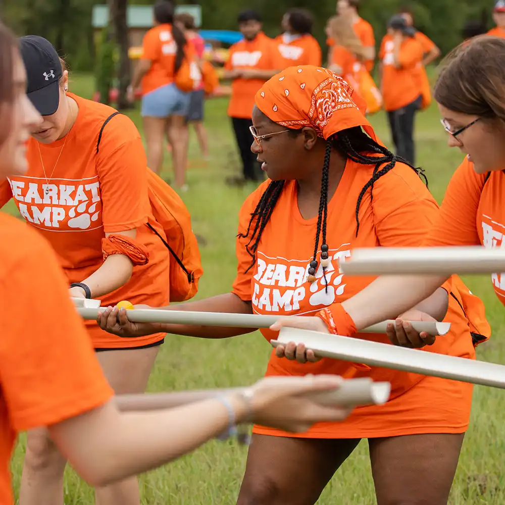 Students wearing bright orange “Bearkat Camp” shirts are participating in an outdoor activity maneuvering a small ball down a handheld chute with several students standing in the background.