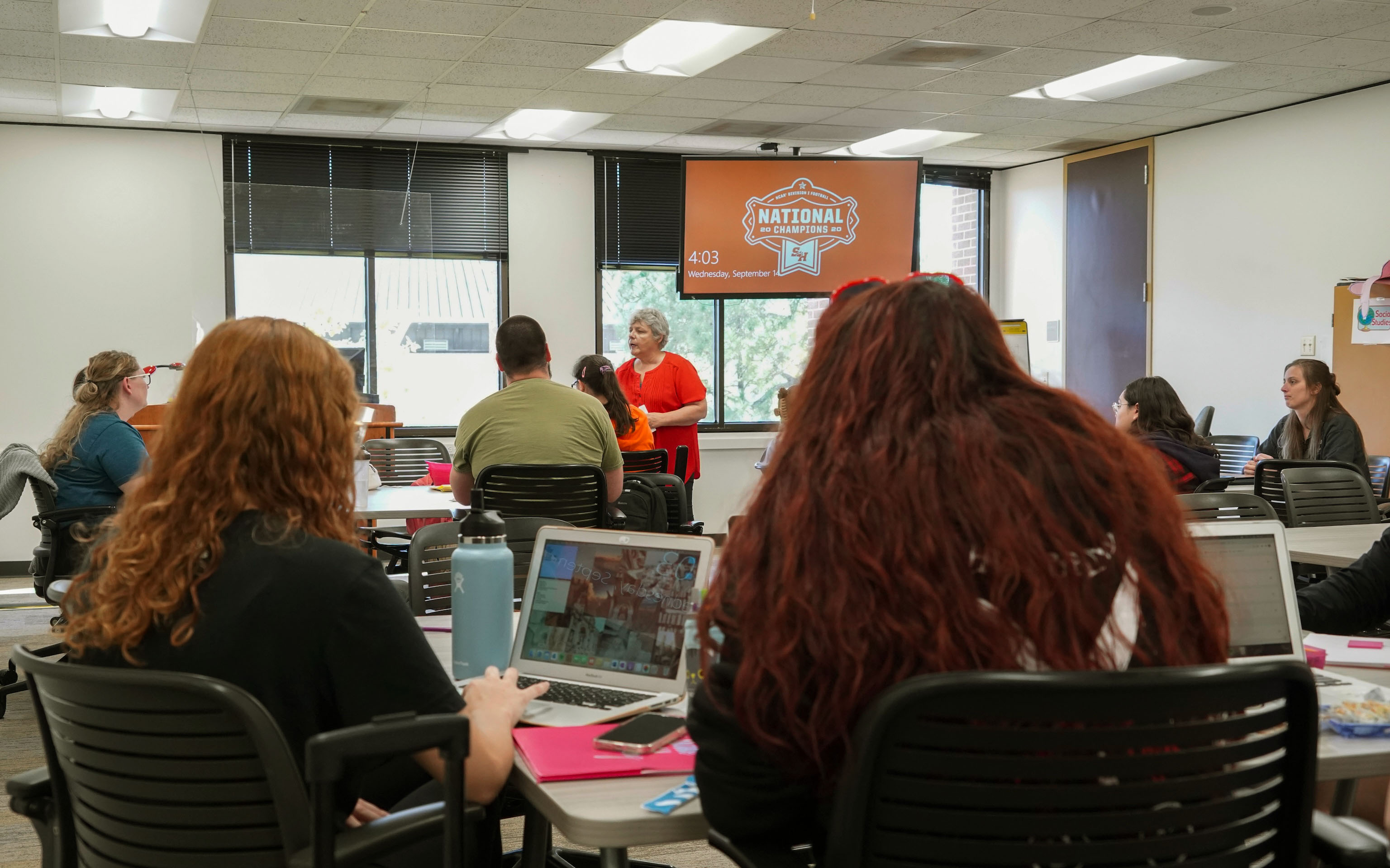 ​​College classroom with students at desks, instructor at the front of the room giving instruction​ 