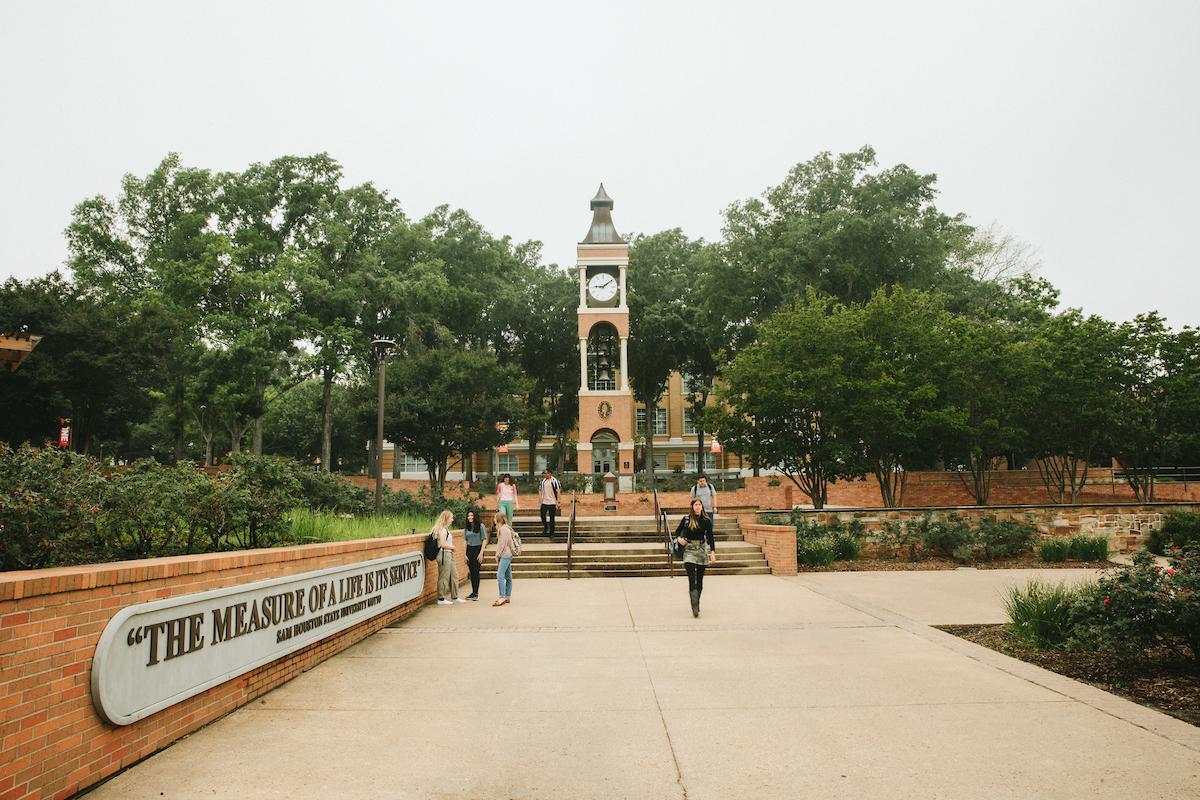 ​​Frank Parker Yard with the SHSU motto on a curved brick wall and the clock tower in the background as students gather.​ 