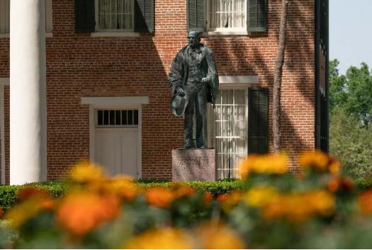 ​​Bronze statue of Sam Houston outside Austin Hall, surrounded by a garden with bright orange and yellow flowers.​ 