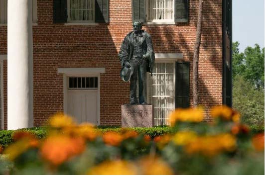 ​​Bronze statue of Sam Houston outside Austin Hall, surrounded by a garden with bright orange and yellow flowers.​ 
