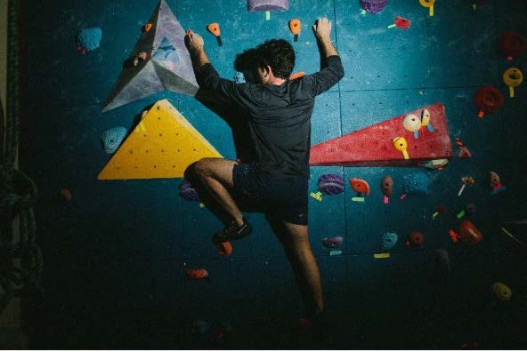 ​​A rock climber pauses on a blue climbing wall, studying the route among various colored holds to decide his next agile move.​ 