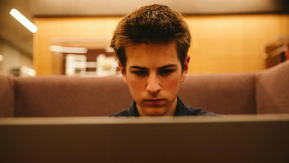 ​​An image of a future student sitting in a cozy booth focusing intently on a laptop as he applies for housing at Sam Houston State University. The student has short brown hair, a navy-blue shirt, and a focused expression. The background features soft lighting and wooden textures, creating a warm and inviting atmosphere. ​ 