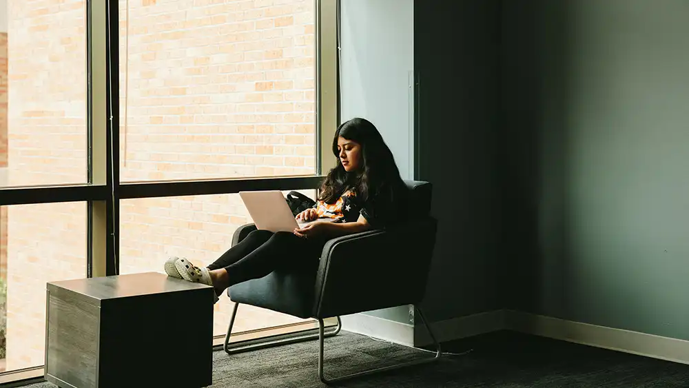 ​​An image depicting a student sitting in a modern lounge area. The student is seated in a dark, cushioned chair by a large window, letting in natural light. The student, wearing a black T-shirt with orange and white accents and white Crocs, is using a laptop. A small, dark coffee table sits in front of the student. The lounge area features a muted color palette, including dark carpet, gray walls, and contemporary furniture. The overall atmosphere is calm and relaxed.​ 