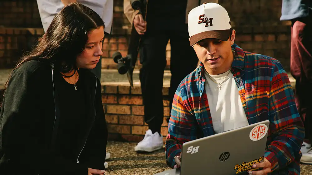 ​​Two college students sit outside on a sunny day, looking at a laptop together. The student on the right wears a Sam Houston State University cap, colorful plaid shirt, and jeans, and is showing something on the screen to the other student. The student on the left wears all black and listens attentively. The laptop is decorated with SHSU-related stickers. Brick steps and other students with skateboards are visible in the background.​ 