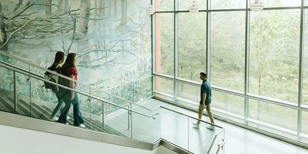 ​​A close-up and wide-angle image of Piney Woods Hall, a modern residence hall located on the campus of Sam Houston State University. The building features a mix of contemporary architectural elements, including large windows, gray metal paneling, and an eye-catching, bright orange overhang at the entrance. The overhang prominently displays the words “PINEY WOODS HALL” in large white block letters. The brick exterior of the building is visible in the background, with rectangular windows evenly spaced across the façade. The photo captures various angles of the building’s entrance, its signage, and parts of the building’s upper floors.​ 